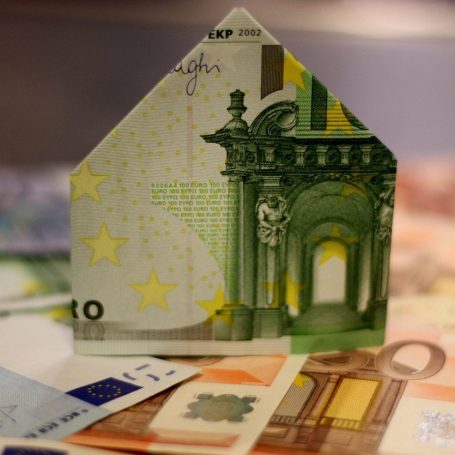 Ein Geldschein, gefaltet in Form eines Hauses steht auf weiteren Geldscheinen.