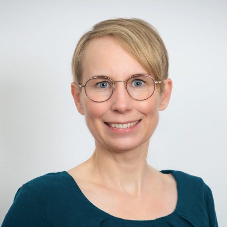 Porträt einer lächelnden Frau mit blonden Haaren und Brille, in einem blauen Oberteil.