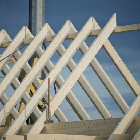 Dachstuhl aus schrägen Holzbalken unter blauem Himmel, Baustelle im Hintergrund.